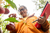 A smiling gardener examines vegetables while holding a tablet and pen in a lush garden, illustrating the integration of technology into modern farming and vegetable growing