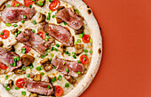 Top view of a gourmet pizza topped with roast beef, porcini mushrooms, cherry tomatoes, green onions and mozzarella cheese, beautifully presented on a warm colour background.