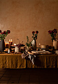 An elegantly laid dinner table with candles, fresh flowers and a variety of appetisers against a warm, textured background.
