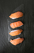 Four pieces of fresh salmon sushi, neatly arranged on a textured black stone surface. The bright orange colour of the salmon contrasts with the dark background and emphasises the elegance.