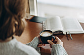 A relaxing scene with a person holding a steaming cup of coffee while an open book lies on a wooden table to capture a peaceful moment of coffee break