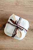 Top view of an elegantly wrapped bento birthday cake, tied with a brown satin ribbon and a wooden spoon, against a rustic wooden background.