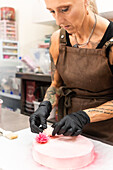 A skilful craftsman wearing an apron and gloves decorates a pink pastry with delicate floral decorations, demonstrating the meticulous work of Italian bakers in the production of elegant desserts
