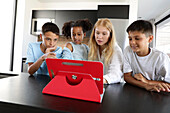 Three children and a young woman gather around a red table and engage in a fun and educational activity. The bright, modern kitchen emphasises the communal atmosphere.