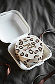 Stylish bento birthday cake with leopard print and gold lettering, neatly presented in a white takeaway box on a textured grey background.