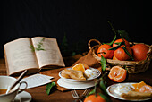 Citrusy crêpes with creamy yoghurt are served in a cosy setting, with a basket of fresh oranges and green leaves adding a lively touch. An open old book emphasises the rustic charm of the dark wooden table.