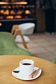 A steaming cup of coffee sits on a wooden table in a cosy café. Blurred chairs and decorations can be seen in the background, adding to the warm and inviting atmosphere