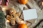 Cosy Christmas scene with a smiling gingerbread man, candy canes, a letter, oranges and fairy lights on a textured background with a blank card, perfect for festive greetings