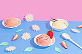 Bright summer picture with bowls of strawberry and vanilla ice cream against a vivid pink and blue background, decorated with daisies, strawberry slices and leaves.