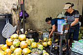 Two women process coconuts in an urban environment. One expertly opens the coconuts with a machete, while the other prepares them for sale in bottles. A busy scene of the tropical fruit trade.