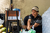 A woman fills bottles with fresh coconut water. She smiles as she works in a casual outdoor setting, surrounded by coconuts and bottles.