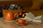 A warm autumn scene with an orange teapot and cup on a wooden table, surrounded by burning candles. An open book in the background creates a cosy, inviting atmosphere.
