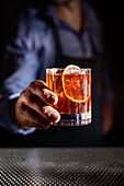 Close-up of an old-fashioned whiskey cocktail held by a hand over a textured bar counter A slice of orange garnishes the drink, contrasting beautifully with its rich amber colour