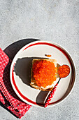 Viewed from above, a healthy organic red caviar made from trout fish is served on a toasted slice of bread, presented on a white plate with a red rim and a dotted napkin under natural sunlight.