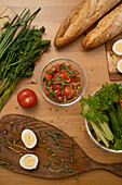 Blick von oben auf frische Zutaten für einen Salat, darunter Kräuter, Tomatensalat, gekochte Eier, Salat, Baguette und grüne Zwiebeln, schön auf einer Holzfläche arrangiert.
