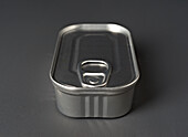 Detailed view of a metal food tin with a pull tab on the lid. The can stands on a smooth, dark grey surface that highlights its shiny metallic texture.