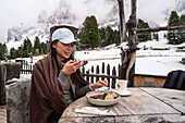 An Asian traveller enjoys a meal in the restaurant of the Geisleralm amidst the breathtaking snow-capped Dolomites. The rustic wooden surroundings offer a tranquil mountain experience