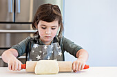 A child in an apron with a star pattern rolls out dough in a concentrated manner. The setting is a bright kitchen where creativity and culinary skills are emphasised in a cheerful home environment.