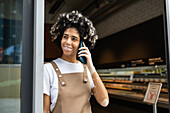 A café employee with vitiligo in a brown apron stands at the entrance and smiles while talking on the phone The atmosphere is casual and friendly and captures a typical working day