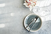Top view of a minimalist dining room. A cracked grey plate with fork and knife, complemented by crystal glasses and a white orchid on a textured surface.