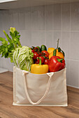 A linen sack filled with fresh vegetables, including peppers, celery and lettuce, lies on a wooden worktop. A symbol of healthy, sustainable living.