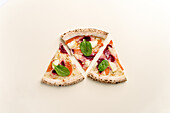Top view of three gourmet pizza slices with beetroot, creamy sauce, mozzarella and a fresh basil leaf, presented against a plain background