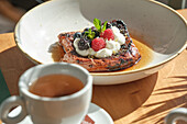 Close-up of French toast topped with fresh blackberries, raspberries, cream and mint, drizzled with syrup A cup of tea completes the cosy snack situation