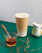 Ein hohes Glas Milchtee, begleitet von einem Glas Honig mit einem Holzlöffel und einer Keramikkanne, arrangiert auf einem strukturierten Hintergrund mit dekorativen Zweigen