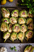 Fresh artichoke halves on a baking tray, surrounded by lemons, herbs and garlic, form a vibrant culinary display, ready for cooking.