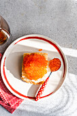 Top view of a bright, tempting plate of organic red trout caviar, served on toast with a red polka dot spoon, against a textured grey background.