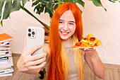 Eine Person mit langem, leuchtendem Haar macht ein fröhliches Selfie, während sie zu Hause ein Stück Pizza genießt Ein Stapel Bücher und Grünpflanzen schaffen eine gemütliche Atmosphäre
