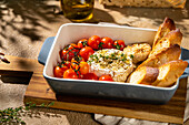 A baked Camembert cheese paired with roasted cherry tomatoes, garlic and crispy baguette slices in a ceramic dish. Enjoy the flavours and texture of this gourmet delight.