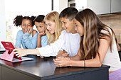 A young woman and four children gather around a tablet, busy and smiling in a modern kitchen. The lively group enjoys a shared digital experience that creates an entertaining and educational atmosphere.