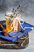 A jar of golden honey, decorated with lavender flowers and tied with a rope, stands on a rustic wooden board A wooden honey spoon and a pair of scissors accompany the arrangement