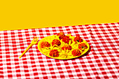A lively arrangement on a red and white chequered cloth with a yellow plate with bright yellow flowers and red blossoms, complemented by a matching yellow fork.