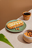 Rustic breakfast set-up with almond butter and banana toast on a green plate, next to a brown ceramic coffee mug and a bowl of almonds on a light-coloured wooden table.