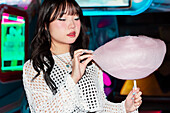 Asian Gen Z girl enjoys candyfloss in a lively arcade The colourful lights illuminate the playful atmosphere and capture youthful joy and modern fun