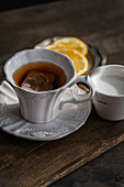 An elegant teacup made of white porcelain with an embossed pattern, filled with English tea and a tea bag. The cup is accompanied by fresh lemon slices on a saucer and a small milk jug.