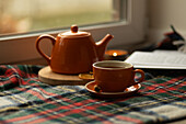 A warm setting with an orange teapot and cup on a chequered blanket near a window. A lit candle and an open book add to the cosy, inviting atmosphere.