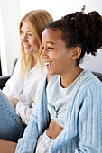A cheerful woman and a girl laugh together while sitting on a couch in a bright living room. The candid moment captures a genuine connection and shows happiness and togetherness.