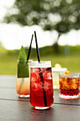 A colourful selection of refreshing cocktails on a table The focus is on a red drink with ice and a black straw in front of a blurred outdoor background