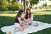 A lesbian couple enjoy a picnic in a lush park. They chat and pour each other wine to celebrate their love and togetherness in a cheerful setting