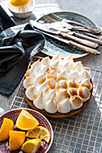 A homemade lemon cake cools on a wire rack, accompanied by orange and lemon slices on a decorative plate. Forks and knives lie next to it on elegant crockery.