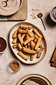 Top view of a ceramic plate with sugary churros next to bowls of chocolate sauce and cinnamon against a background of rustic linen and wooden kitchenware.