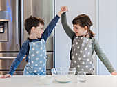 Two young siblings in star-patterned aprons enthusiastically raise their hands in the air while baking in a modern kitchen, capturing cheerful teamwork and family cohesion.