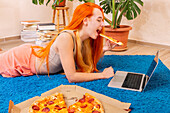 A person with flowing hair enjoys a slice of pizza while relaxing with a laptop on a blue carpet.