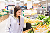 A young woman with a plait and casual hoodie selects fresh vegetables in a brightly lit supermarket aisle. She looks down at the produce.
