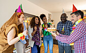 A cheerful group of people are celebrating a birthday in a house. They are wearing colourful party hats and strings of flowers and gather eagerly around a cake with lit candles.