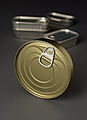 Close-up of several metal cans, highlighting a closed can in the foreground, its lid and pull-tab in the centre, and other open cans in the background against a grey surface.
