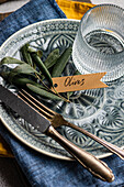 A sophisticated table setting with olive leaves on a ceramic plate with a shield, a glass and fine dining utensils.